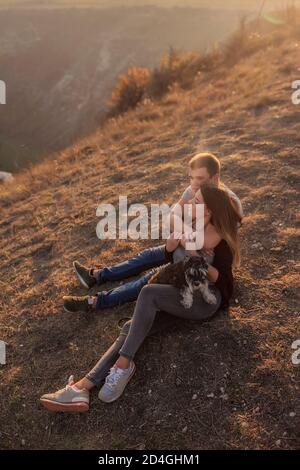 Un jeune homme embrasse une belle fille de derrière, le couple admirer le coucher du soleil depuis le sommet de la colline. Une famille heureuse voyage avec un chien schnauzer. Banque D'Images