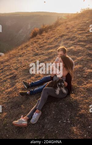 Un jeune homme embrasse une belle fille de derrière, le couple admirer le coucher du soleil depuis le sommet de la colline. Une famille heureuse voyage avec un chien schnauzer. Banque D'Images