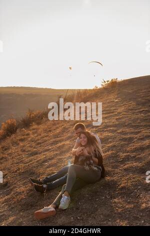 Un jeune homme embrasse une belle fille de derrière, le couple admirer le coucher du soleil depuis le sommet de la colline. Une famille heureuse voyage avec un chien schnauzer. Banque D'Images