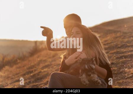 Un jeune homme embrasse une belle fille de derrière, le couple admirer le coucher du soleil depuis le sommet de la colline. Une famille heureuse voyage avec un chien schnauzer. Banque D'Images