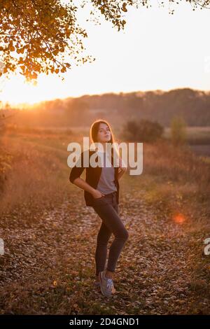 Une fille moderne, belle, dans une veste bordeaux, jeans foncés, se tient dans un champ, les cheveux blonds flirtent dans le vent contre le fond d'un automne Banque D'Images