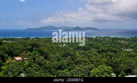 Vue aérienne de la magnifique côte nord de l'île de la Digue, Seychelles avec forêt tropicale, village, littoral et île voisine de Praslin. Banque D'Images