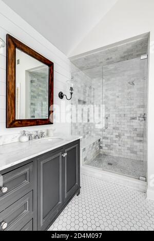 Luxueuse salle de bains rénovée avec meuble-lavabo gris, miroir rustique à cadre en bois, douche avec porte vitrée et carrelage en marbre. Banque D'Images