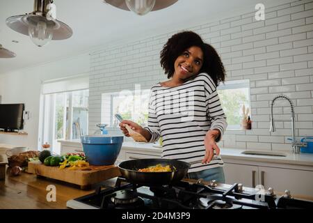 Femme ethnique souriante discutant sur un smartphone tout en tenant une spatule pour préparer un déjeuner dans une casserole dans une cuisine moderne. Banque D'Images