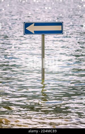 Flèche de la route du panneau d'affichage à gauche partiellement submergée par les eaux d'inondation. Un panneau routier se trouve au milieu d'un lac d'eau. Banque D'Images