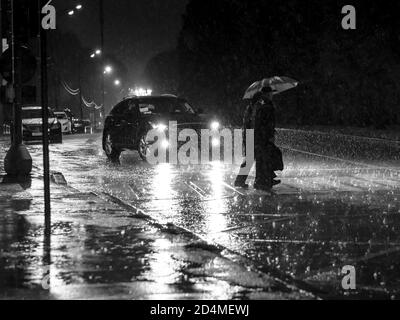 Vue sur une traversée piétonne de la ville la nuit pendant une lourde averse. Silhouettes de personnes avec parasols dans les phares de la voiture. Personnes Banque D'Images