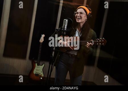Portrait d'une jeune femme, artiste féminine jouant ukulele et chantant dans un microphone à condensateur tout en enregistrant une chanson dans un studio professionnel Banque D'Images