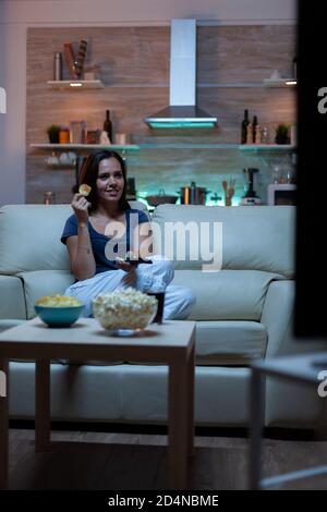 Femme appréciant la soirée regarder des séries tv à la maison assis sur un canapé confortable habillé en pyjama. Excitée et amusante maison seule dame mangeant des en-cas et de jus de fruits sur un canapé confortable dans la salle de séjour. Banque D'Images