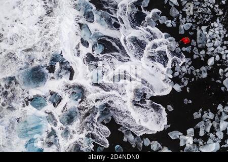 Homme vêtu de rouge qui photographie les vagues entre les rochers de glace dans la célèbre plage noire islandaise. Plage noire, sorcière diamants de glace paysage à votre calendrier Banque D'Images