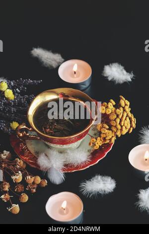 Thé rouge blanc vintage et or avec des herbes vertes sur une table noire sombre. Camomille séchée, lavande, fleurs jaunes, bougies allumées Banque D'Images