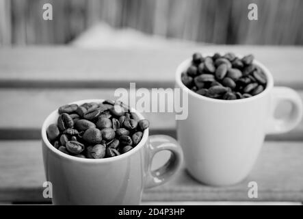 Gros plan en niveaux de gris de deux tasses complètes avec café rôti haricots sur une table en bois Banque D'Images