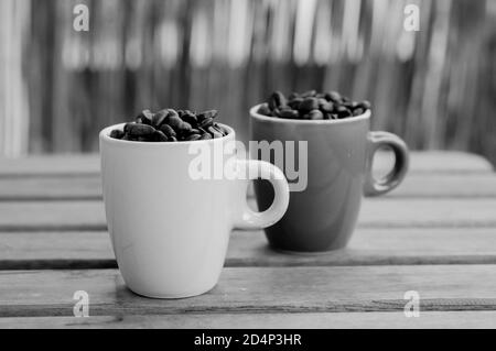 Gros plan en niveaux de gris de deux tasses complètes avec café rôti haricots sur une table en bois Banque D'Images