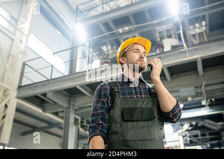 Jeune ingénieur confiant en casque de protection et vêtements de travail utilisant un walkie talkie Banque D'Images