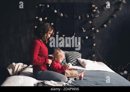 Mère tenant bébé fille de 1-2 ans portant des chandails tricotés rouges sur le décor de Noël dans la chambre. Saison des vacances d'hiver. Maternité. Noël. Banque D'Images