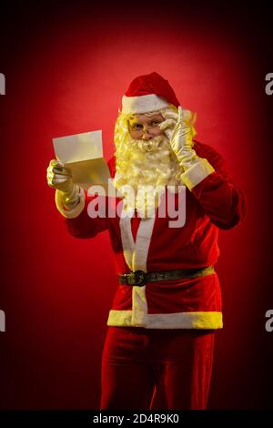 L'acteur masculin habillé comme le Père Noël tient une feuille de Papier dans ses mains une lettre de Noël lit et pose sur un arrière-plan rouge foncé Banque D'Images