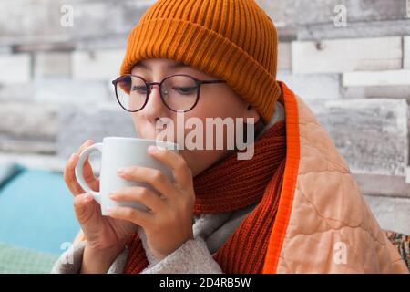 Une fillette mal de gorge s'assoit sur un canapé sous une couverture et boit du thé chaud. Elle porte un foulard et un chapeau. Banque D'Images