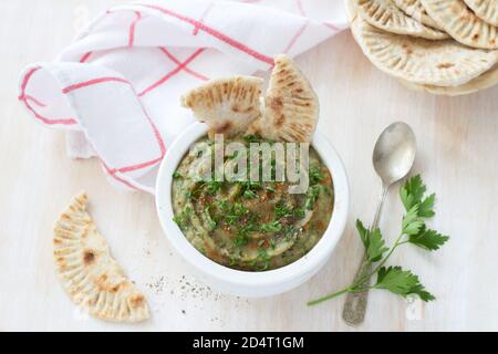 Trempez l'aubergine dans un bol en céramique blanc avec persil et pain plat à grains entiers sur fond de bois clair Banque D'Images