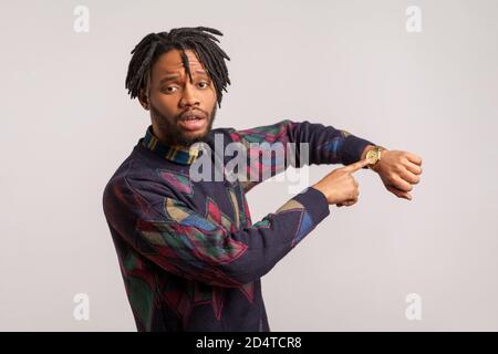 Homme africain nerveux et anxieux avec des dreadlocks et une barbe pointant du doigt sur sa montre-bracelet, vérifiant l'heure avec impatience, étant en retard, échéance. Salle de studi Banque D'Images