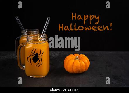 Deux pots carrés en verre avec araignée noire. Mocktail de citrouille orange sans alcool et pailles en verre sur une surface texturée noire. La citrouille entière est près des bocaux. Banque D'Images