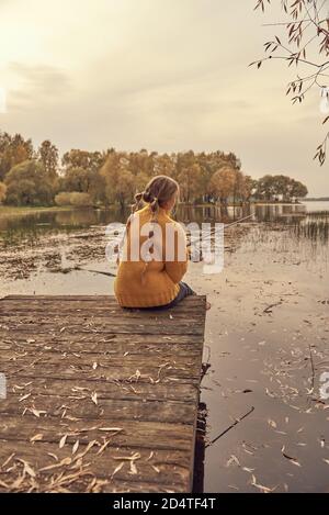 Une fille dans un chandail tricoté jaune est assise sur un pont en bois au-dessus du lac et attrape du poisson. Banque D'Images