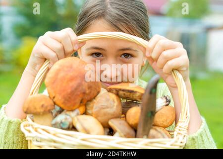 Divers champignons crus dans un panier en osier sorcière caucasienne fille tient dans ses mains. Elle regarde de dessous la poignée du panier.le couteau est dans le panier. Champignons porcini, champignons de bouleau, champignons boletus à chapeau orange. Concentrez-vous sur le visage des filles. Mise au point sélective. Banque D'Images