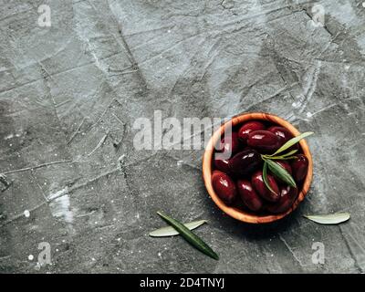 Olives Kalamata sur fond gris texturé. Olives rouges kalamata dans un bol en bois d'olive avec feuilles d'olives vertes fraîches, vue sur le dessus ou plat. Copier l'espace pour le texte ou la conception. Banque D'Images