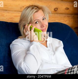 Une blonde d'âge moyen dans une maison de campagne en bois parle au téléphone avec un sourire. Travailler à domicile. Travail à distance. Banque D'Images