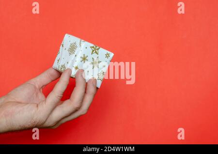 Cadeau de Noël enveloppé dans la main d'un homme. Une main tient une petite boîte avec un cadeau sur fond rouge. Concept de nouvel an et de Noël Banque D'Images