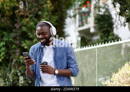 Marche dans la rue, détente, loisirs. Jeune Afro-américain dans des écouteurs écouter de la musique sur un smartphone à l'aide de l'application musique. Portrait de sourire Banque D'Images