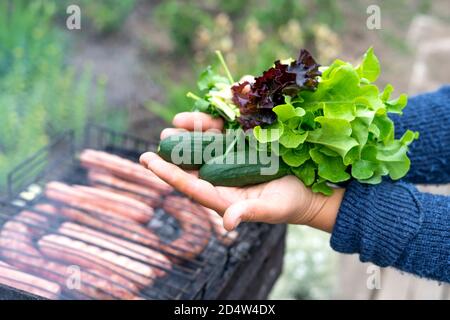 Les mains des femmes tiennent les ingrédients de la salade : concombres, laitue, basilic. Pique-nique. Les saucisses sont cuites sur le gril. Mise au point sélective. Vue rapprochée. Banque D'Images
