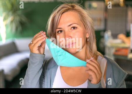Photo rapprochée d'une femme caucasienne aux cheveux blonds posée sur un masque médical au bureau. Elle ajuste son masque médical avec ses mains et souriant. Banque D'Images