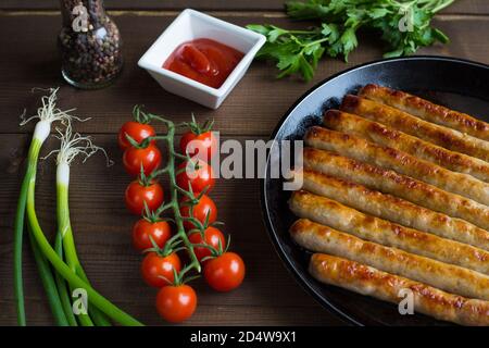 Des saucisses grillées, entières et délicieuses sont posées sur la poêle, qui repose sur un fond en bois de planches. À proximité se trouvent une branche de tomates cerises fraîches, Banque D'Images