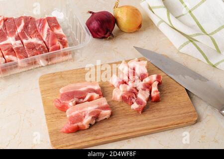 Couper le ventre frais de porc cru en petits morceaux sur une planche à découper en bois avec un couteau tranchant au-dessus d'une table de cuisine. Viande dans un récipient en plastique jetable Banque D'Images