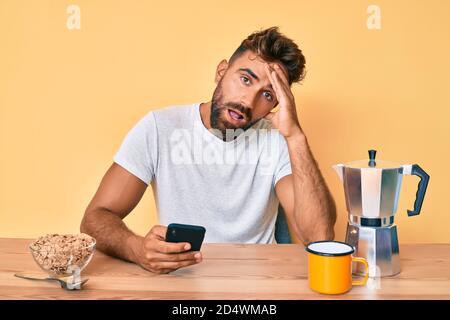 Jeune hispanique homme assis sur la table prenant le petit déjeuner et utilisant smartphone stressé et frustré de la main sur la tête, surpris et fâché visage Banque D'Images