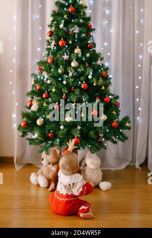Bébé dans un costume tricoté drôle est en recherche pour un Décoration de Noël sur un arbre de Noël Banque D'Images