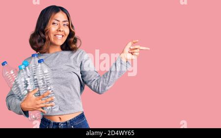 Jeune belle femme de race mixte tenant des bouteilles en plastique de recyclage souriant pointez avec votre main et votre doigt sur le côté Banque D'Images
