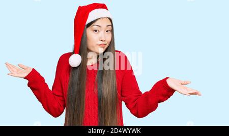 Jeune femme chinoise portant un chapeau de noël expression sans indice et confuse avec les bras et les mains soulevées. Doute concept. Banque D'Images