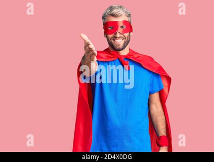 Jeune homme blond portant un super héros crème souriante amical offrant poignée de main comme accueil et accueil. Affaires réussies. Banque D'Images