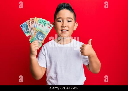 Petit garçon hispanique enfant tenant des dollars australiens souriant heureux et positif, pouce vers le haut faisant excellent et signe d'approbation Banque D'Images
