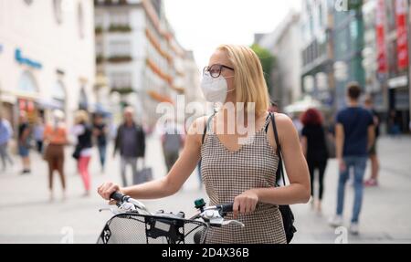 Femme marchant à vélo dans la rue piétonne de la ville portant un masque médical en public pour empêcher la propagation du virus corona. Nouvelle normale pendant Banque D'Images