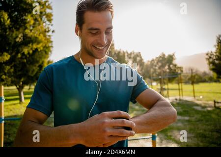 Jeune athlète masculin vérifiant Smartwatch souriant portant des écouteurs pendant faites de l'exercice dans un parc extérieur Banque D'Images