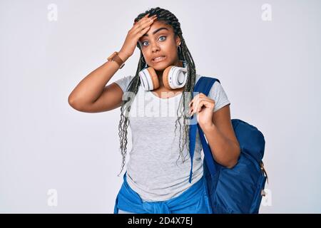 Jeune femme afro-américaine avec des tresses portant un sac à dos étudiant et des écouteurs stressés et frustrés de la main sur la tête, surpris et furieux visage Banque D'Images