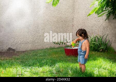 Photo au niveau des yeux d'une petite fille mignonne avec un arrosage peut être jouet sous l'eau qui éclabousse dans un jardin Banque D'Images