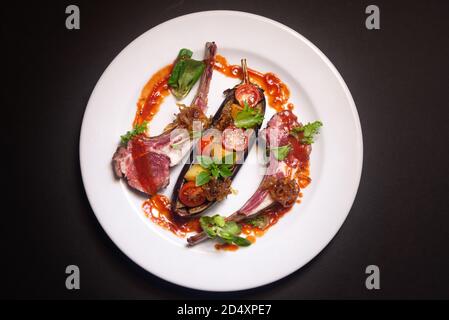 Côtes d'agneau rare degré de préparation de la viande, aubergine cuite au four, quartiers de tomates cerises, oignons caramélisés, feuilles de basilic, micro-légumes, sauce rouge Banque D'Images