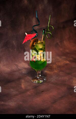 Grand verre de jus à base de kiwi avec morceaux de fruits, glace, paille et parasol sur fond gris foncé. Gros plan. Banque D'Images