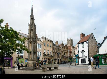 Centre-ville de Glastonbury et Market Cross entièrement restauré. Les foules qui se frapissent avec deux jeunes hommes s'embuent d'un accueil. Magasins locaux visibles. Banque D'Images