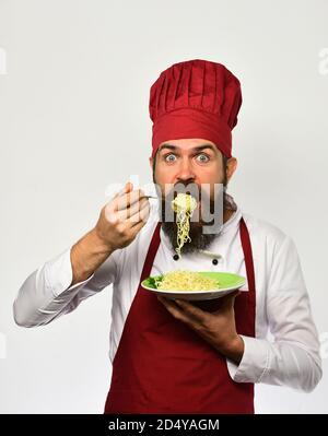 Le chef mange des nouilles italiennes ou asiatiques. Concept de cuisine de restaurant. Cuire avec le visage surprise dans l'uniforme bordeaux tient la fourchette et l'assiette. L'homme avec la barbe tient un plat savoureux sur fond blanc. Banque D'Images