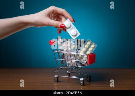 La main d'une femme avec une manucure rouge met des pilules dans un petit chariot. Arrière-plan bleu. Le concept du coronavirus, des affaires et des produits pharmaceutiques. Banque D'Images