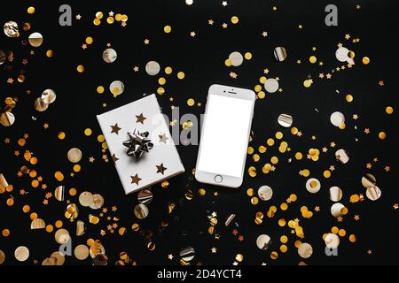 Téléphone cellulaire avec écran blanc vierge ou espace de copie ou maquette et à côté d'une boîte avec un cadeau sur fond noir avec confetti d'or. Historique conceptuel de la vente des fêtes. Banque D'Images