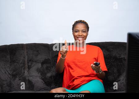 femme africaine assise sur un canapé et tenant une télévision La télécommande pointe vers le téléviseur pour sourire et donner un pouce haut Banque D'Images